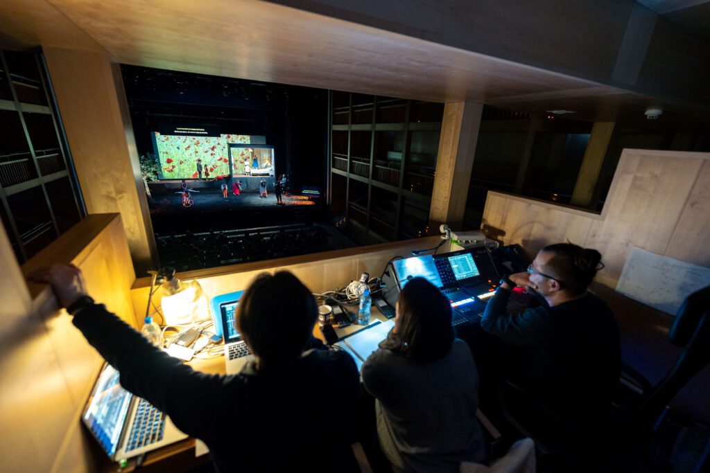 widok z reżyserki na scenę, widok na 3 osoby siedzące tyłem do obiektywu przy konsolecie w perspektywie scena i kolorowy spektakl