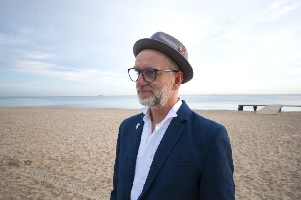 artysta Grzegorz Nawrocki, mężczyzna w garniturze, z krótką brodą, w okularach i kapeluszu. Zdjęcie na plaży, W perspektywie może i niebo.